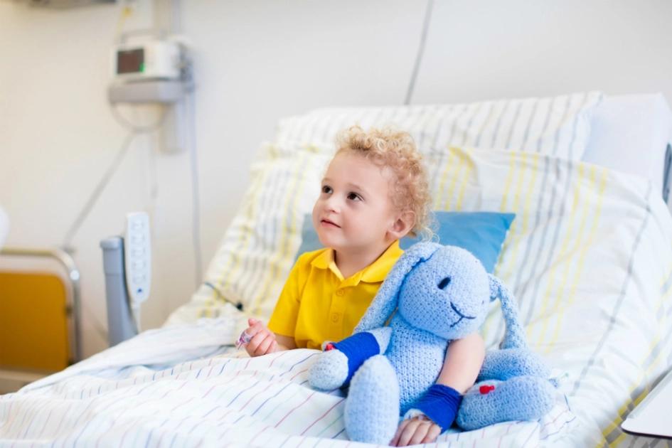 liten pojke med gul tröja sitter med sin nalle i en sjukhussäng och ler mot någon utanför bilden.jpg