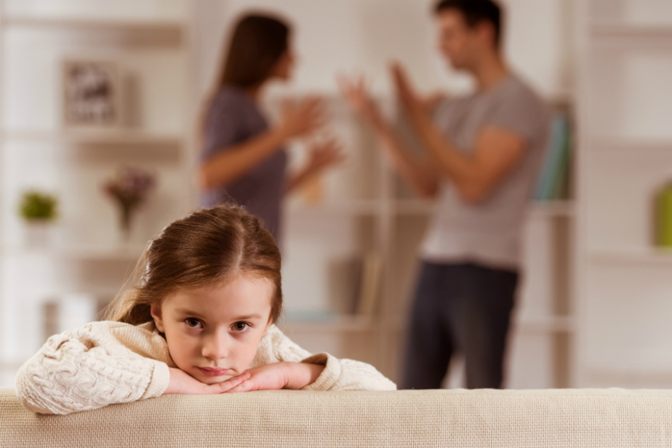 flicka sitter och tittar i kameran över en soffkant medan föräldrar bråkar i bakgrunden.jpg