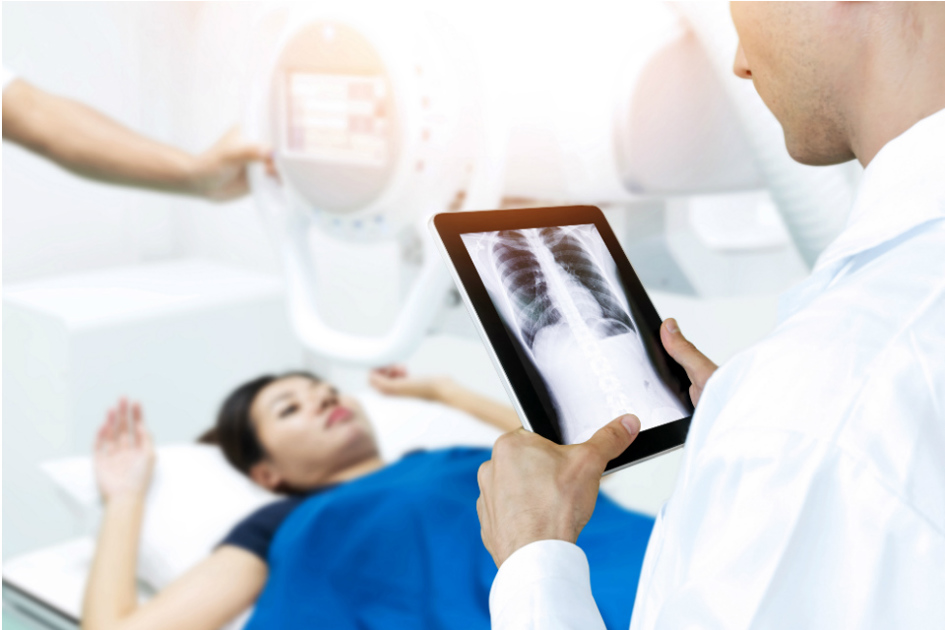 kvinna i bakgrunden ligger på vårdbädd; man i förgrunden tittar på hennes röntgenplåt på en platta.jpg