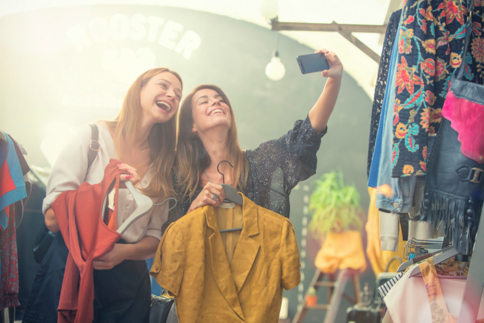 två tjejer i en klädbutik håller i varsitt plagg och den ena av dem tar en selfie på sig och sin kompis.jpg