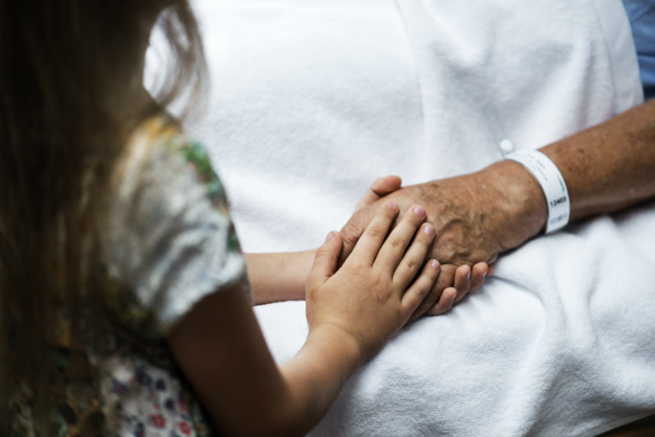 ryggen på en liten flicka som håller en äldre man i sjukhussäng i handen.jpg